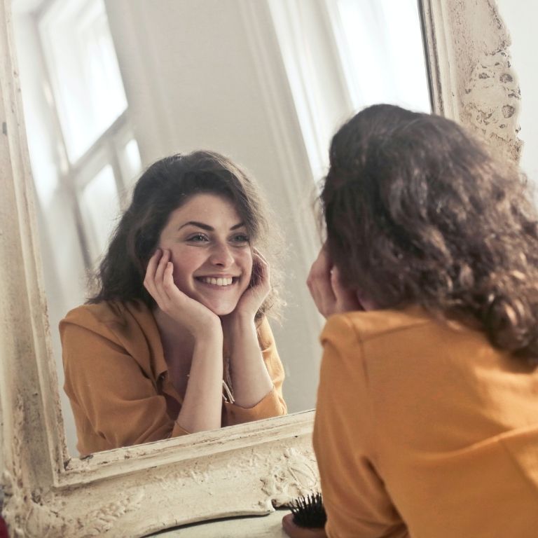 Woman looking at herself in the mirror smiling. Accepting who she is and embracing her flaws. She is taking responsibility for her life.